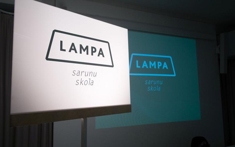 Izgaismota zīme ar tekstu "LAMPA sarunu skola" telpā ar projekciju, kurā redzams tas pats teksts. Atmosfēra ir izglītojoša un profesionāla.