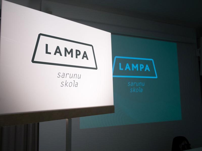 Izgaismota zīme ar tekstu "LAMPA sarunu skola" telpā ar projekciju, kurā redzams tas pats teksts. Atmosfēra ir izglītojoša un profesionāla.