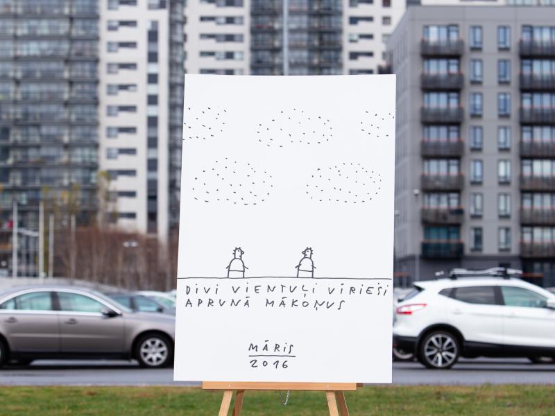 Liels, ar roku zīmēts plakāts uz molberta attēlo divas figūras zem punktētiem mākoņiem ar tekstu "Divi vientuļi vīrieši aprunā mākoņus" apakšā. Plakāts novietots ārā uz augstu, modernu ēku fona.