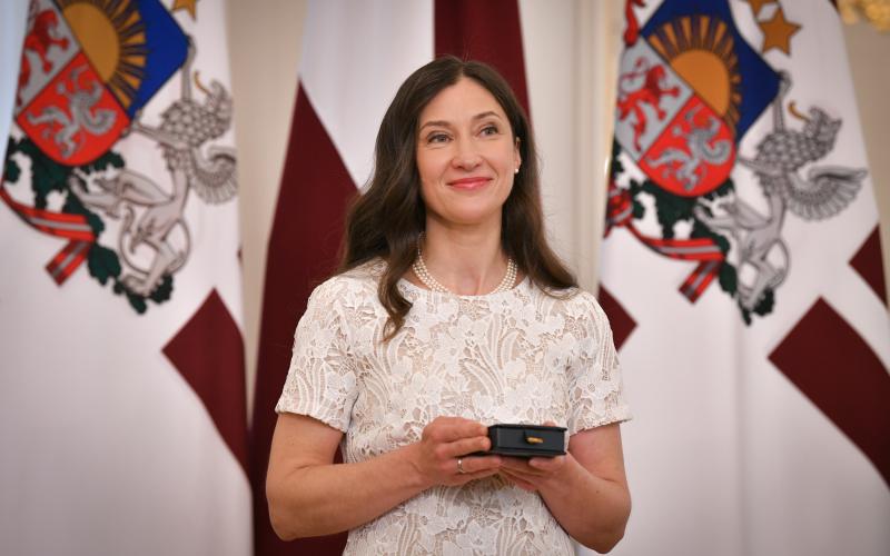 Ieva Morica, tērpta baltā mežģīņu kleitā, smaida, turot rokās nelielu kastīti ar saņemto Triju Zvaigžņu ordeni. Viņa stāv Latvijas karoga un Latvijas Valsts prezidenta standarta priekšā.