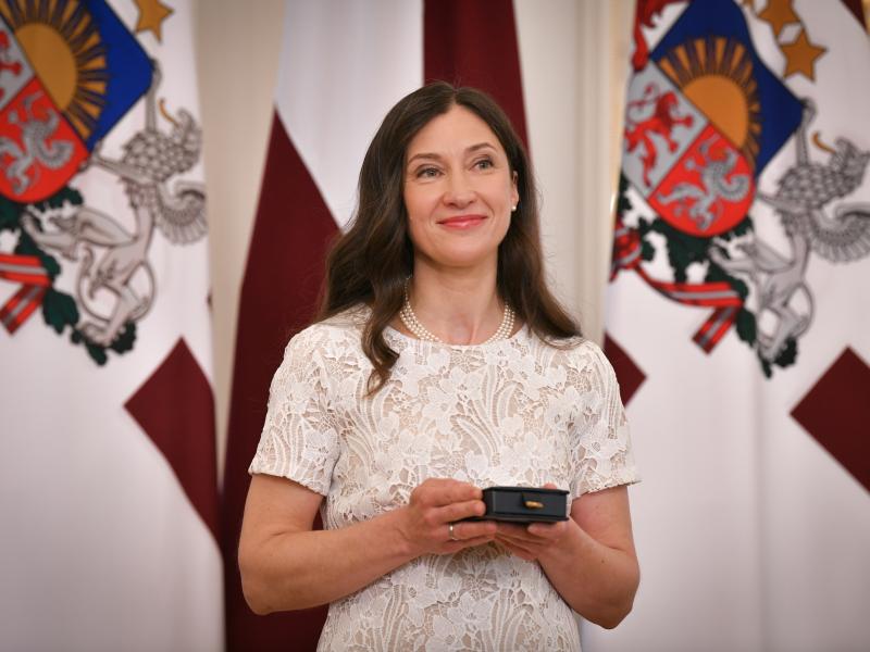 Ieva Morica, tērpta baltā mežģīņu kleitā, smaida, turot rokās nelielu kastīti ar saņemto Triju Zvaigžņu ordeni. Viņa stāv Latvijas karoga un Latvijas Valsts prezidenta standarta priekšā.