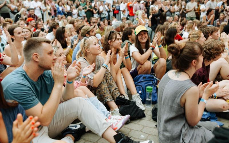 Dažādu cilvēku pūlis sēž ārā uz ietves, aplaudē un smaida, radot svētku un priecīgu atmosfēru festivālā.