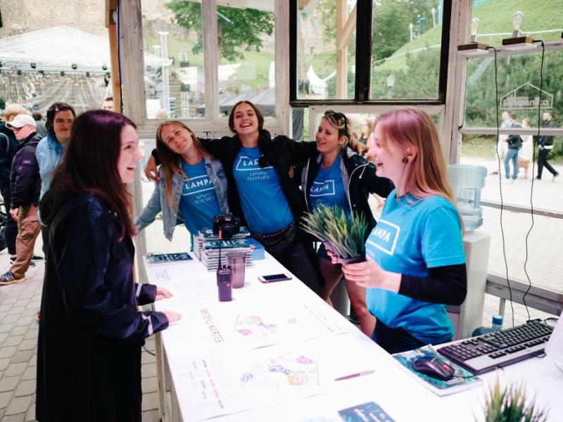 Četri smaidoši Sarunu festivāls LAMPA brīvprātīgie zilos kreklos stāv aiz informācijas stenda, sirsnīgi sarunājoties ar apmeklētāju. Atmosfēra ir rosīga un atrodas brīvā dabā.