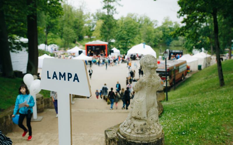 Dzīvīga parka aina ar norādes zīmi "LAMPA", akmens statuju un pūļiem, kas sapulcējušies pie baltām teltīm un skatuves.