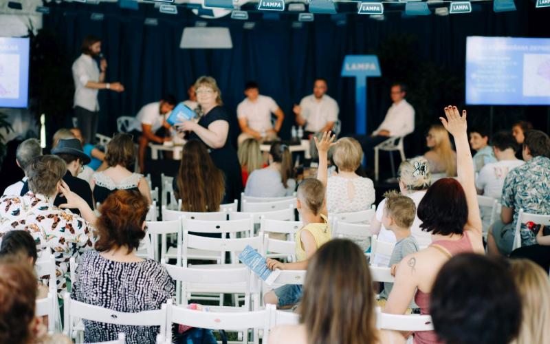 Pasākuma auditorija, kurā cilvēki paceļ rokas, sarunājoties ar runātāju grupu uz skatuves gaišā, pārpildītā telpā. Atmosfēra ir dzīvīga.