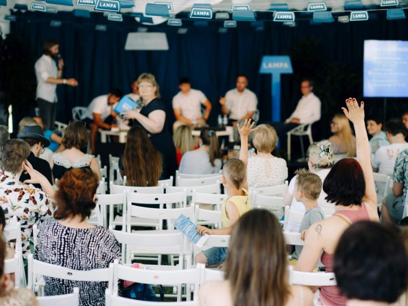 Pasākuma auditorija, kurā cilvēki paceļ rokas, sarunājoties ar runātāju grupu uz skatuves gaišā, pārpildītā telpā. Atmosfēra ir dzīvīga.