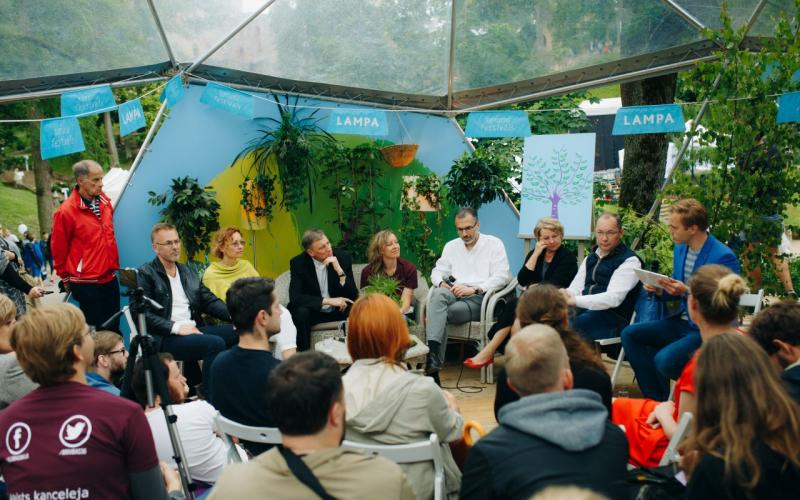 Astoņu cilvēku paneļdiskusija notiek zem caurspīdīgas telts, ko ieskauj zaļumi. Priekšplānā sēž uzmanīga auditorija.