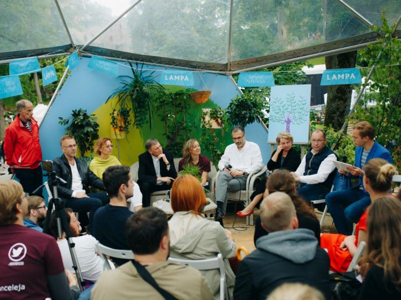 Astoņu cilvēku paneļdiskusija notiek zem caurspīdīgas telts, ko ieskauj zaļumi. Priekšplānā sēž uzmanīga auditorija.