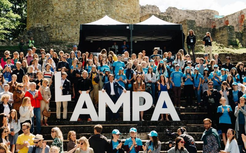Uz pakāpieniem pulcējās dažādu cilvēku pūlis, pozējot ar lieliem burtiem "LAMPA". Gaiša vasaras diena, dzīvespriecīga atmosfēra, fonā pilsdrupas.