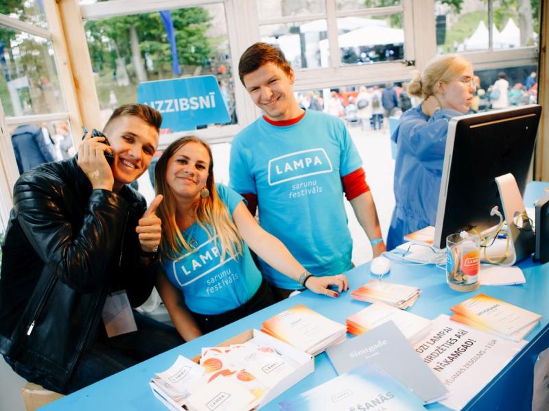 Trīs smaidoši cilvēki pie festivāla stenda ģērbušies zilos "LAMPA" kreklos, uz galda atrodas bukleti. Viens cilvēks tur rokās telefonu, rādot īkšķi uz augšu. Noskaņojums ir dzīvīgs un viesmīlīgs.