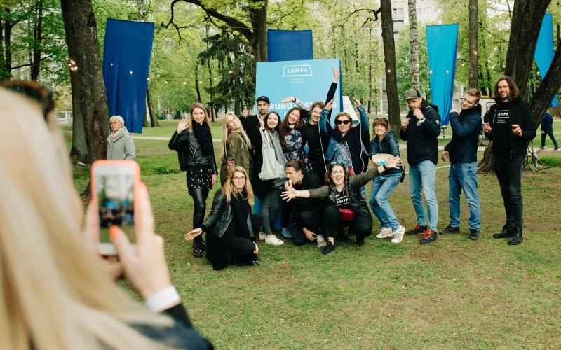Priecīgu cilvēku grupa pozē fotogrāfijai zaļā parkā, daži paceļ rokas. Priekšplānā esoša persona iemūžina mirkli ar tālruni. Fonā redzami zili baneri un koki, radot svētku atmosfēru.