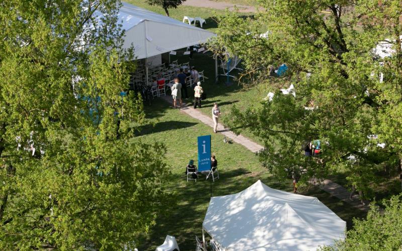 Skats no gaisa uz saulainu pasākumu brīvā dabā, kurā cilvēki pastaigājas pa takām starp baltām teltīm, ko noēno zaļi koki, radot dzīvīgu, aicinošu atmosfēru.