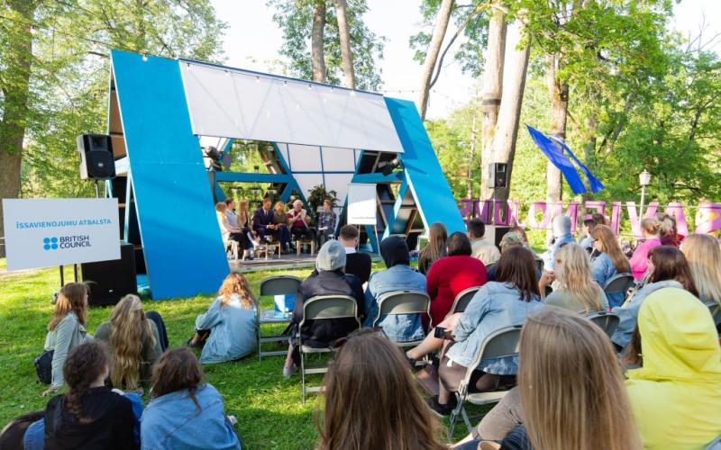 Āra paneļdiskusija ar daudzveidīgu auditoriju parkā. Moderna zila skatuve, kas novietota starp kokiem, rada dzīvīgu un saistošu atmosfēru.