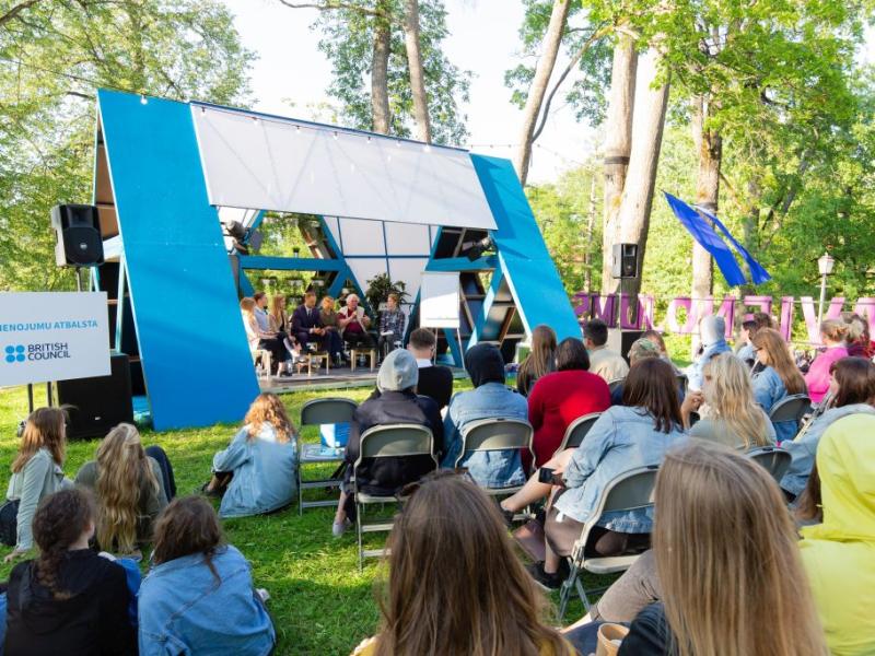 Āra paneļdiskusija ar daudzveidīgu auditoriju parkā. Moderna zila skatuve, kas novietota starp kokiem, rada dzīvīgu un saistošu atmosfēru.
