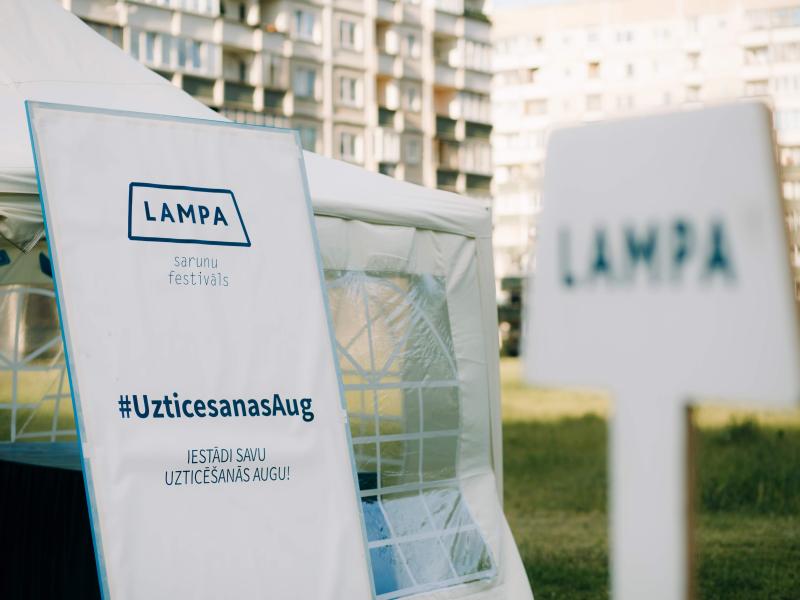 "Festivāla telts ar 'LAMPA' baneri un mirkļbirku #UzticesanasAug pilsētas parka vidē. Dzīvokļu ēkas izplūdušas fonā, saulaina atmosfēra."