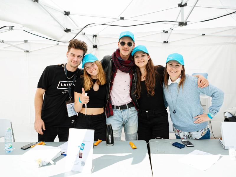 Piecu smaidīgu cilvēku grupa, kas stāv kopā zem baltas telts, ģērbušies ikdienas apģērbā un zilās cepurēs, izstarojot dzīvespriecīgu un draudzīgu atmosfēru.