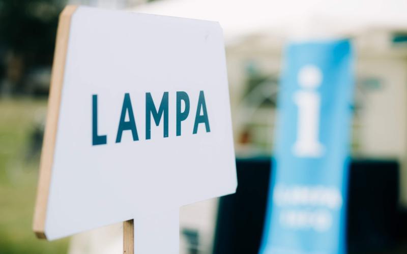 Baltas zīmes tuvplāns ar uzrakstu "LAMPA" trekniem melniem burtiem uz izplūduša āra fona ar zilu informācijas baneri, radot informatīvu toni.