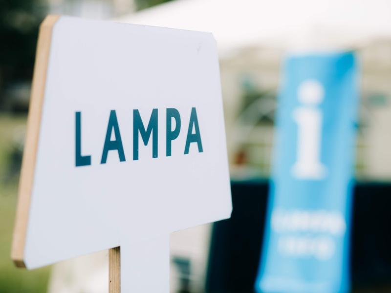 Baltas zīmes tuvplāns ar uzrakstu "LAMPA" trekniem melniem burtiem uz izplūduša āra fona ar zilu informācijas baneri, radot informatīvu toni.