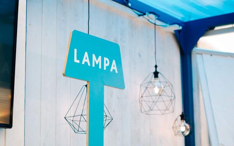 Zila zīme ar baltu uzrakstu "LAMPA" uz gaišas koka sienas. Netālu atrodas ģeometriskas piekaramās gaismas, kas rada modernu un gaisīgu atmosfēru.