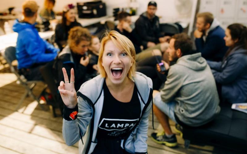 Priekšplānā priecīga sieviete rāda miera zīmi, plati smaidot. Fonā cilvēku grupa mierīgi sēž, sarunājas un ir atslābinājusies.