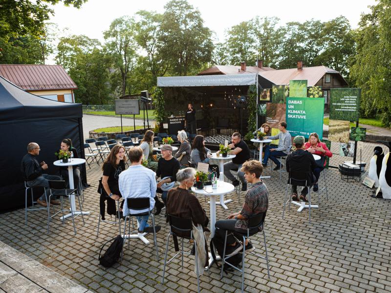 Neformāla pulcēšanās brīvā dabā, kur cilvēki sēž pie nelieliem galdiņiem bruģētā laukumā. Fonā uz skatuves uzstājas runātājs. Apstādījumi un relaksēta atmosfēra.