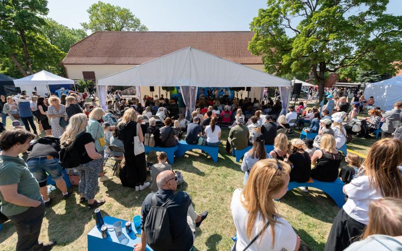 Brīvdabas festivāla aina ar pūli, kas sēž uz ziliem soliem un stāv zem baltas nojumes. Koki un zilas debesis rada dzīvīgu atmosfēru.
