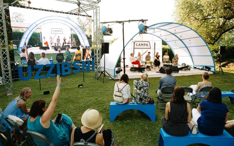 Āra paneļdiskusija festivālā zem izliektas nojumes. Dalībnieki sēž uz soliņiem un komunicē ar runātājiem uz skatuves. Gaiša, dzīvīga atmosfēra.