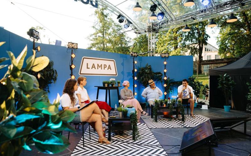 Piecu cilvēki sēž uz skatuves ar uzrakstu "LAMPA" uz zila fona. Viņus ieskauj augi, radot relaksējošu āra atmosfēru.
