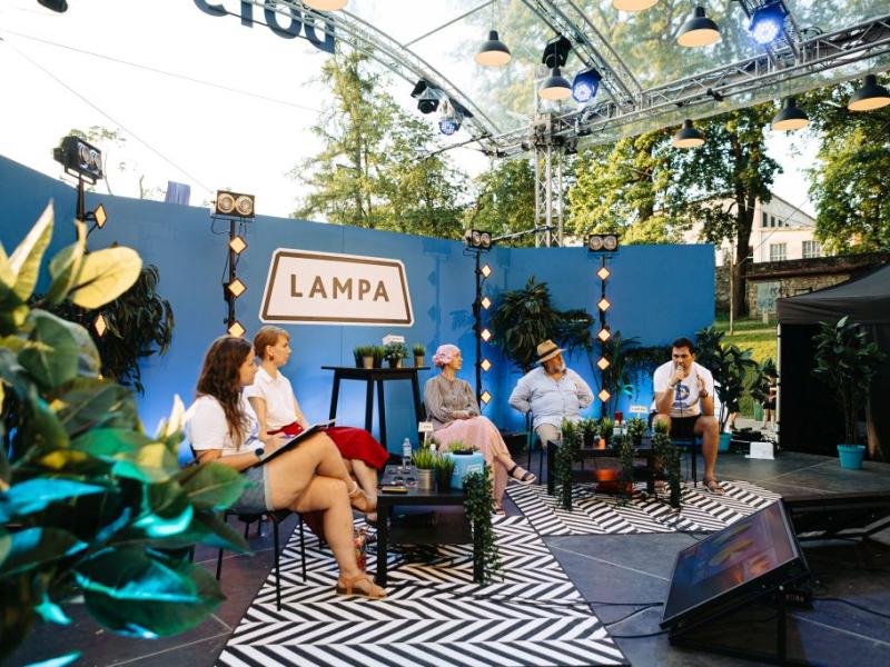 Piecu cilvēki sēž uz skatuves ar uzrakstu "LAMPA" uz zila fona. Viņus ieskauj augi, radot relaksējošu āra atmosfēru.
