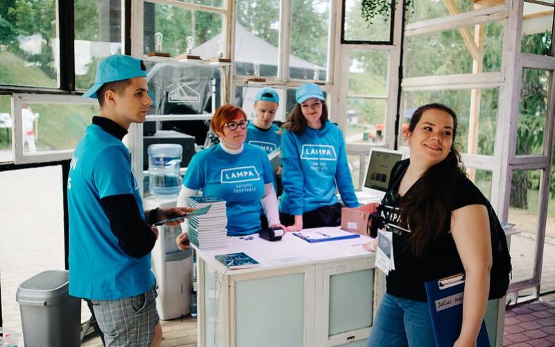 Festivāla brīvprātīgo grupa vienādos zilos formas tērpos stāv pie informācijas stenda. Vide ir gaiša un mājīga.