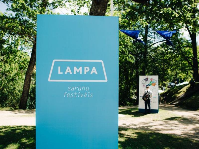 Saulainā parkā stāv liela zila zīme ar uzrakstu "LAMPA sarunu festivāls". Netālu pastaigājas cilvēki, un starp sulīgi zaļiem kokiem plīvo zili karogi, radot svētku atmosfēru.