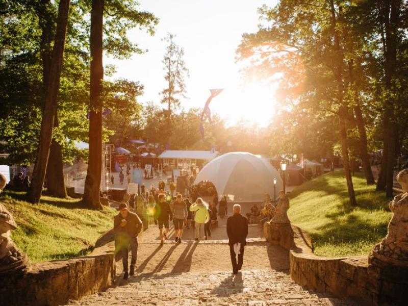 Saulains parka skats ar cilvēkiem, kas iet pa akmens taku kupola formas telts virzienā. Taku ieskauj augsti koki, radot siltu un aicinošu atmosfēru.