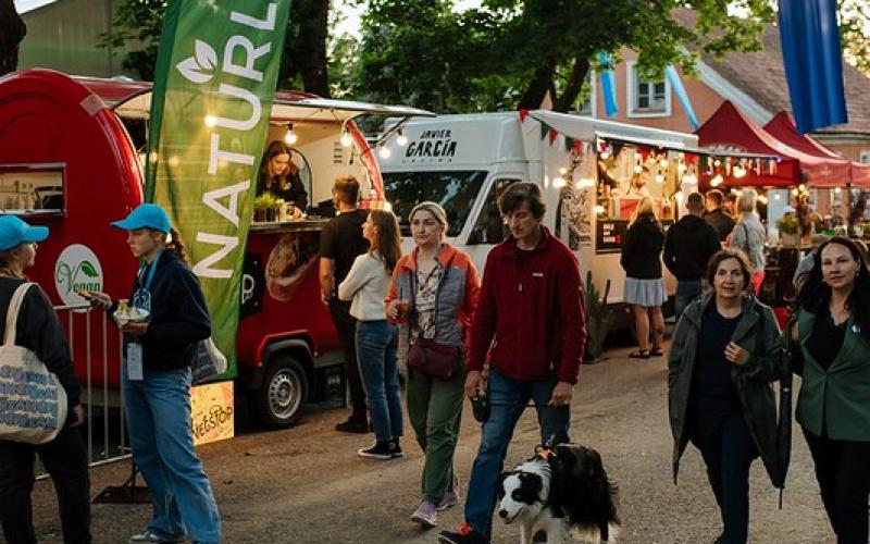 Cilvēki pastaigājas pa festivāla ēdināšanas zonu svētku atmosfērā. Gar taku stāv ēdienu furgoni ar krāsainiem plakātiem un zīmēm.