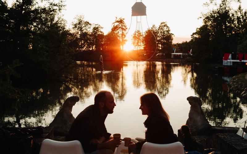 Divi cilvēki sēž uz baltiem krēsliem mierīga ezera krastā, rokās turot krūzes, un viņu silueti kontrastē ar saulrietu.