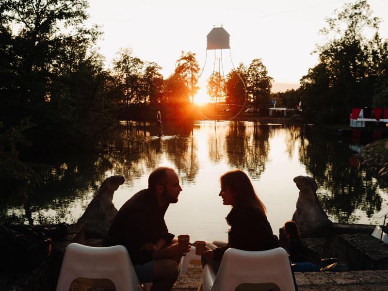 Divi cilvēki sēž uz baltiem krēsliem mierīga ezera krastā, rokās turot krūzes, un viņu silueti kontrastē ar saulrietu.