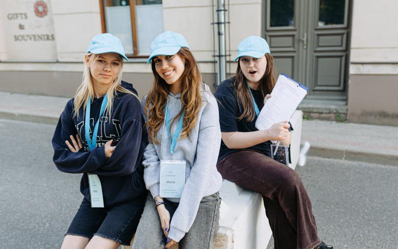 Trīs jaunas sievietes - festivāla brīvprātīgās -zilās cepurēs pozē ikdienišķi. Viena tur rokās planšeti.