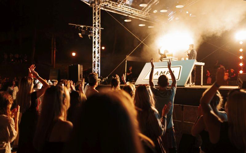 Politiķu disko aina ar gavilējošu pūli, kas vērsts pret spilgti apgaismotu skatuvi, kur uzstājas divi dīdžeji. Atmosfēra ir enerģiska un dzīvespriecīga.