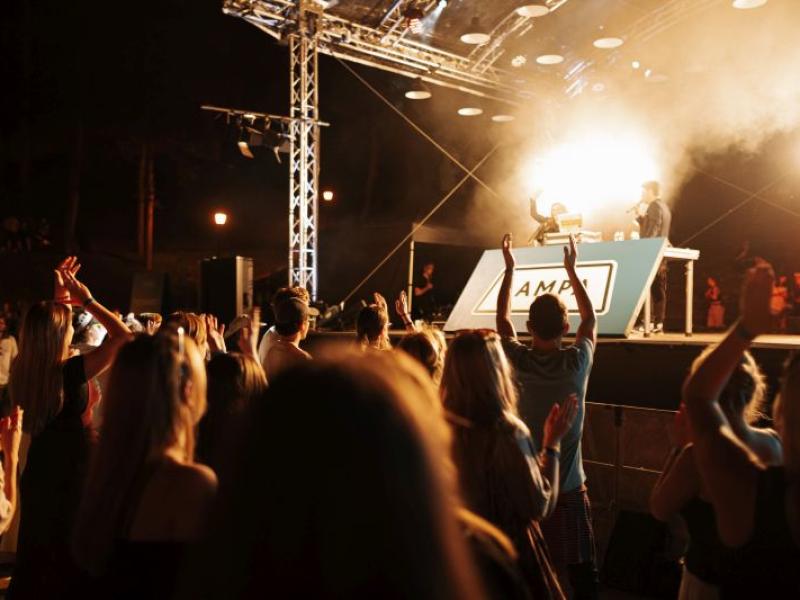 Politiķu disko aina ar gavilējošu pūli, kas vērsts pret spilgti apgaismotu skatuvi, kur uzstājas divi dīdžeji. Atmosfēra ir enerģiska un dzīvespriecīga.