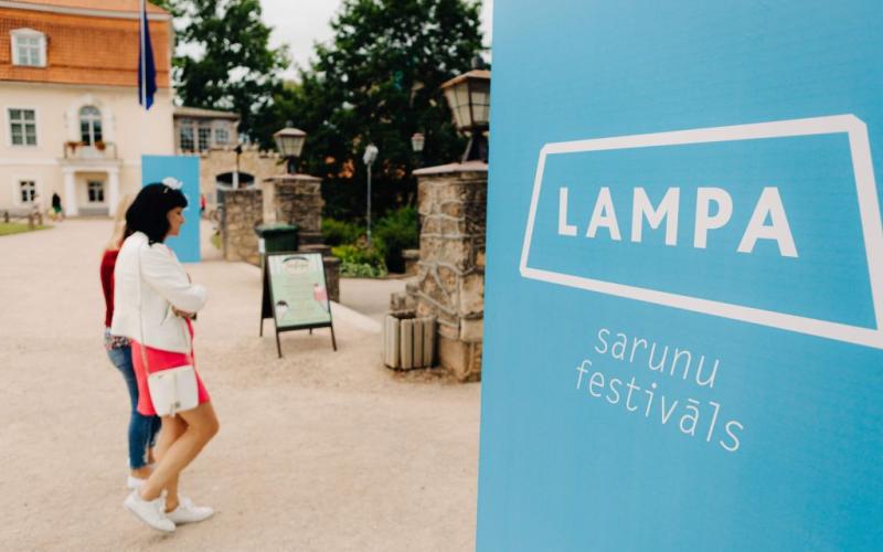 Priekšplānā izceļas zila zīme ar baltu uzrakstu "LAMPA sarunu festivāls". Divas sievietes paiet garām mierīgā āra vidē ar vēsturiskām ēkām.