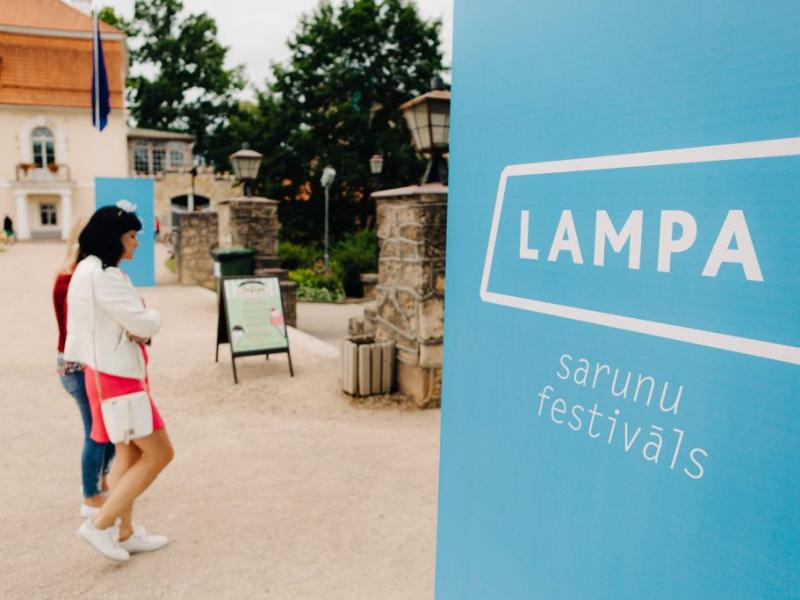 Priekšplānā izceļas zila zīme ar baltu uzrakstu "LAMPA sarunu festivāls". Divas sievietes paiet garām mierīgā āra vidē ar vēsturiskām ēkām.