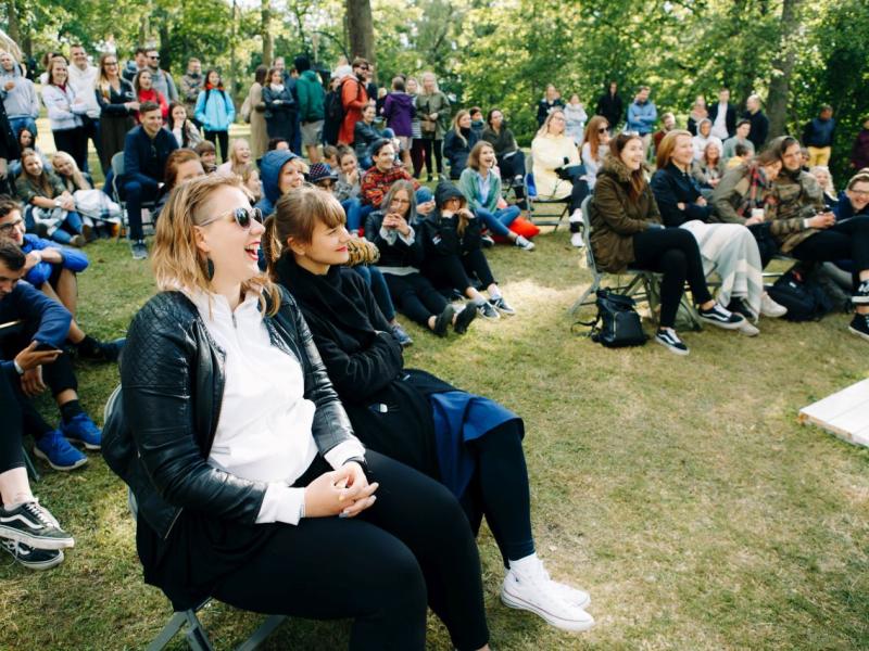 Dzīvīga publika brīvā dabā sēž zālē, smejas un bauda pasākumu. Koki un zaļumi rada mierīgu fonu. Pūlis ir atslābināts un iesaistīts.