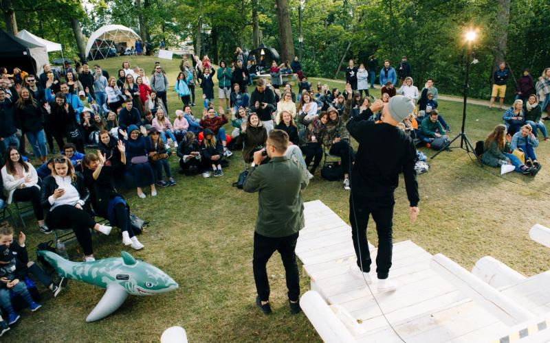 Divi izpildītāji stāv pretī publikai brīvā dabā, sēžot uz zāles, ko ieskauj koki. Mierīgā publika šķiet iesaistīta un dzīvespriecīga, ar festivāla atmosfēru.