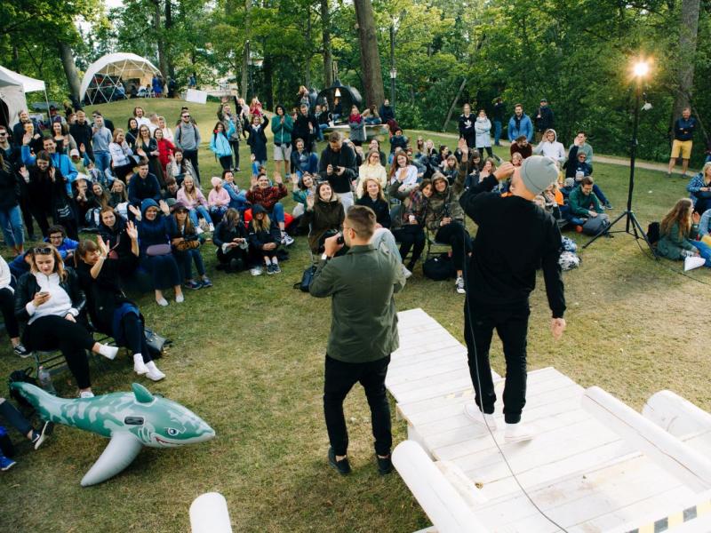 Divi izpildītāji stāv pretī publikai brīvā dabā, sēžot uz zāles, ko ieskauj koki. Mierīgā publika šķiet iesaistīta un dzīvespriecīga, ar festivāla atmosfēru.