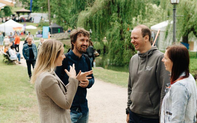 Četri cilvēki stāv uz takas parkā, iesaistoties dzīvīgā sarunā un smejoties. Fonā redzami zaļumi, teltis un cilvēki.