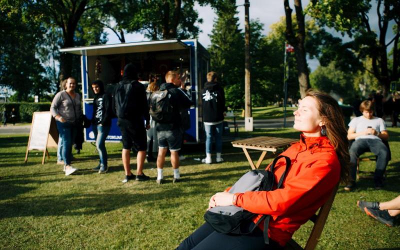 Sieviete sarkanā jakā atpūšas saulainā dienā parkā ar aizvērtām acīm un smaidot. Fonā cilvēki pulcējas pie zila ēdienu furgona starp zaļiem kokiem.