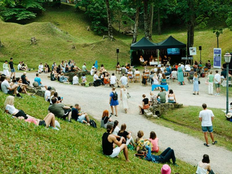 Pulcēšanās brīvā dabā parkā, kur cilvēki sēž uz zāles un krēsliem, vērsti pret melnu telti. Noskaņojums ir mierīgs sulīgi zaļojošu augu vidū.