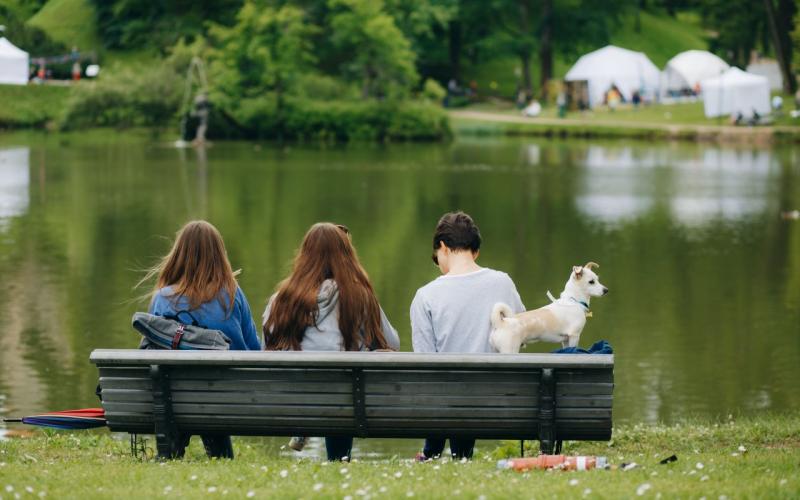 Trīs cilvēki sēž uz soliņa mierīga ezera krastā, fonā koki un teltis. Blakus viņiem stāv mazs suns, radot mierīgu ainu.