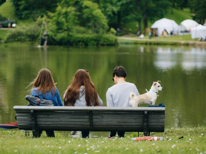 Trīs cilvēki sēž uz soliņa mierīga ezera krastā, fonā koki un teltis. Blakus viņiem stāv mazs suns, radot mierīgu ainu.
