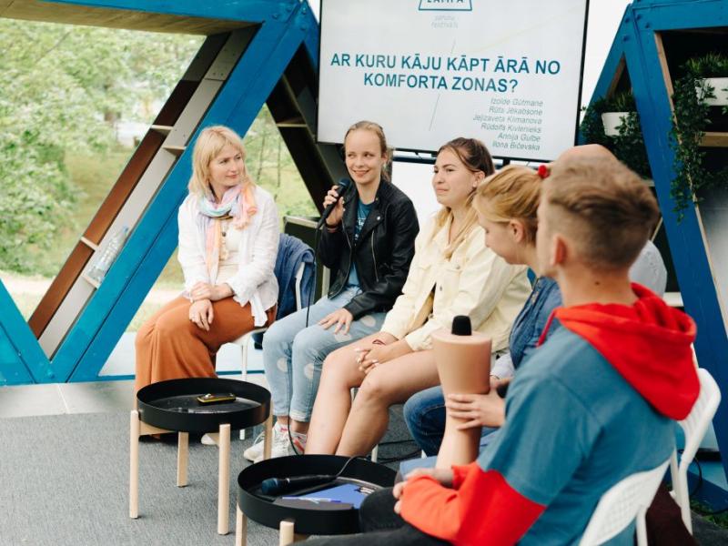 Seši cilvēki sēž puslokā ārā un iesaistās dzīvā diskusijā. Sieviete smaida un tur mikrofonu. Fonā ir uzraksts "Ar kuru kāju kāpt ārā no komforta zonas?".