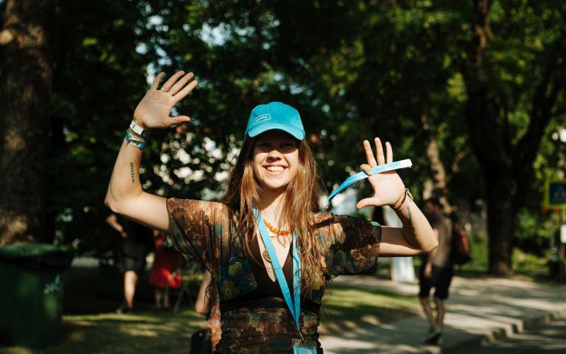 Smaidošs cilvēks zilā cepurē priecīgi paceļ rokas saulainā parkā. Viņam mugurā ir ziedu raksta krekls, kas rada bezrūpīgu noskaņu.